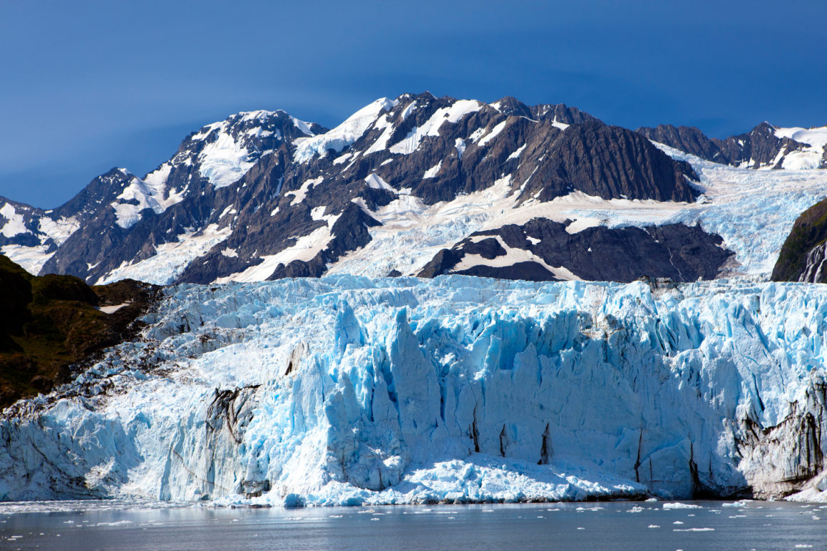 Encosta de uma geleira em tons azulados, com alto paredão de gelo, tendo ao fundo uma cadeira de montanhas rochosas, com pontos cobertos de neve