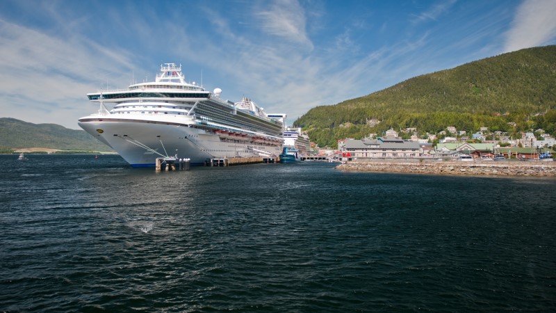 Navio atracado no porto de Ketchikan, no Alasca, tendo a sua frente o mar azul e como paisagem no fundo a cidade e um morro coberto de vegetação verde
