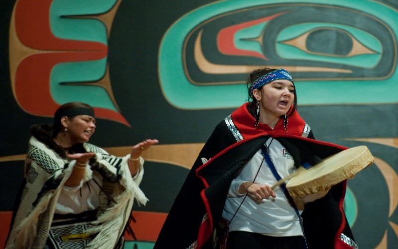 Num cenário de teatro, na cidade de Sitka no Alasca, uma mulher toca um pandeiro com uma baqueta, vestindo uma capa de estilo étnico e uma faixa azul na cabeça, enquanto outra mulher dança e canta, com um poncho branco