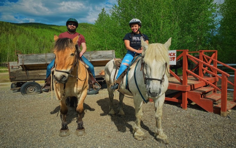 Um homem montado num cavalo marrom e uma mulher montada num cavalo branco pousam para a foto sorrindo, tendo ao fundo uma paisagem de árvores verdes e uma carroça de madeira.
