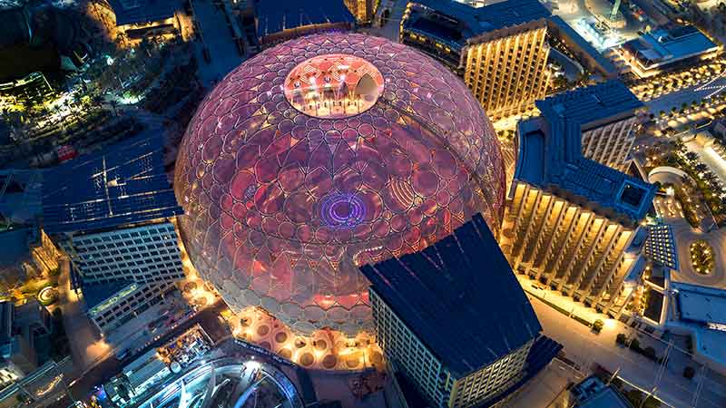 Na Expo 2020 Dubai, vista aérea do Domo Al Wasl iluminado, durante a noite, tendo sua cúpula iluminada por uma projeção rosa