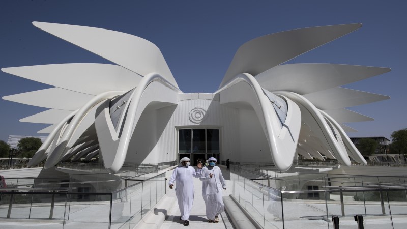 Na Expo 2020 Dubai, dois homens vestidos com roupas típicas dos Emirados Árabes, vestes longas e brancas, com lenços brancos na cabeça, saem do Pavilhão dos Emirados Árabes, uma construção branca com as laterais formadas por estruturas em forma de pena. Projeto do arquiteto espanhol Santiago Calatrava.