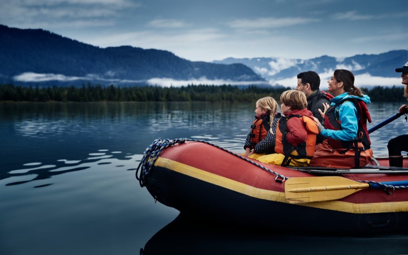 Família composta por um casal de homem e mulher e duas crianças loiras passeiam num bote inflável vermelho, no mar do Alasca. Todos vestem colete salva-vidas.
