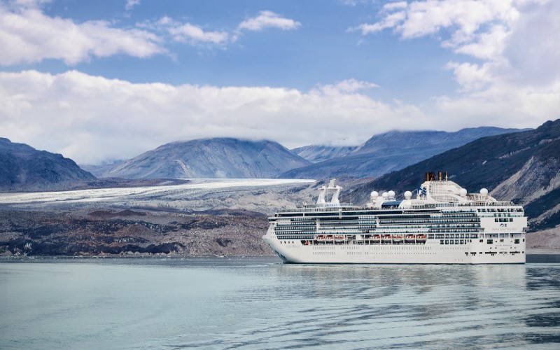 Navio percorre a encosta das geleiras, navegando próximo a uma paisagem rochosa, tendo ao fundo montanhas altas e a geleira