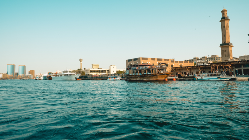 Canal Dubai Creek, no centro de Dubai, com águas azuis e barcos atracados no pier, tendo como fundo as construções antigas da cidade.