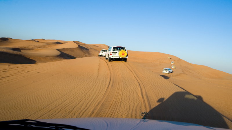 Um comboio de jipes 4x4 percorre as dunas de Dubai, tendo em primeiro plano as areais e ao fundo, céu azul.