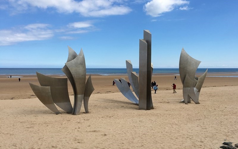 cruzeiro fluvial pela Europa: Praia de Omaha, na Normandia, com três esculturas sobre a areia, pessoas andando e mar ao fundo.