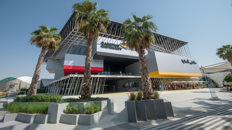 Pavilhão da Alemanha na Expo Dubai, uma alta estrutura quadrada formada por três volumes fechados e perfis metálicos, tendo a frente um jardim de coqueiros.