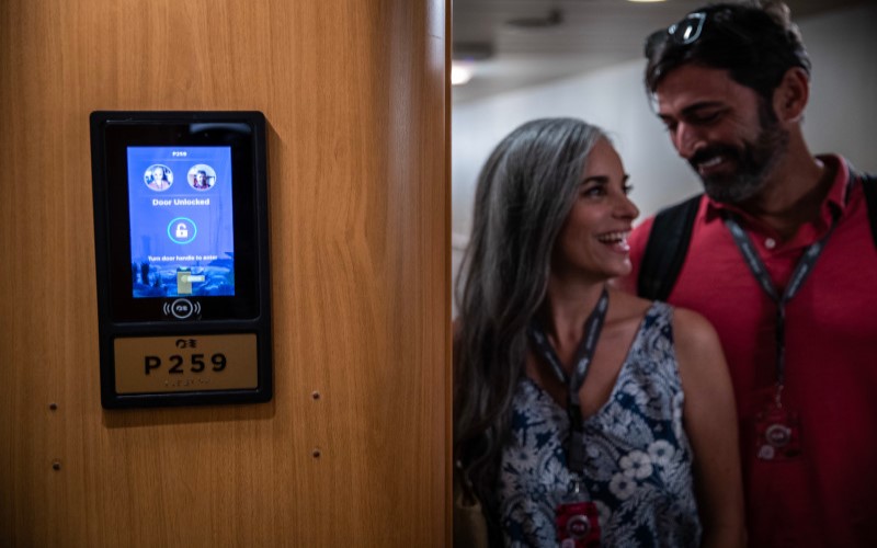 Casal sorrindo se aproxima da porta da cabine, que possui uma tela indicando seus nomes e fotos.