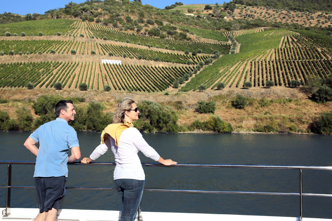 Casal no deck de um navio fluvial admira a paisagem da margem, coberta de vinhedos.