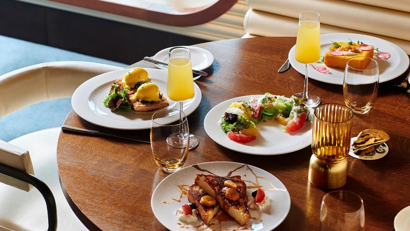 Mesa de madeira redonda do restaurante The Wake no navio Scarlet Lady, da Virgin Voyages, decorado com quatro pratos brancos contendo as refeições.