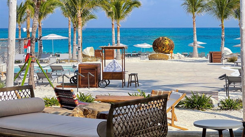 Beach Clube na ilha de Bimini é uma das paradas do Scarlet Lady, da Virgin Voyages, tendo ao fundo o mar e céu azuis e guarda-sóis colocados na areia branca da praia. Em primeiro plano, uma piscina decorada com banquetas, cadeiras de praia e sofás.