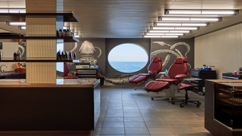 Estúdio de tatuagem do Scarlet Lady, novo navio da Virgin Voyages, decorado com duas cadeiras vermelhas e parede decorada por uma lula gigante, tendo ao centro uma janela redonda com vista para o mar.