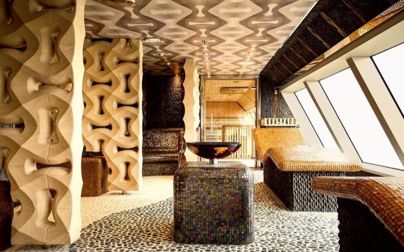 Imagem de um ambiente do Spa Samsara no navio Costa Diadema, com cadeiras espreguiçadeiras revestidas de pastilhas de azulejo, chão com mosaico de pedras e pia de pedra de cor âmbar.
