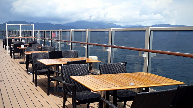 Terraço ao ar livre com vista para o mar, na lateral do navio MSC Seaside, com mesas de madeira e cadeiras, ao lado de um guarda-corpo de vidro.
