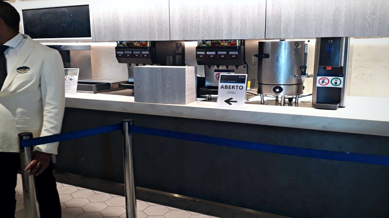 Interior do navio MSC Seaside: estação de bebidas com máquinas de sucos e cafés, tendo a frente uma fita isolando a área e um atendente do navio.