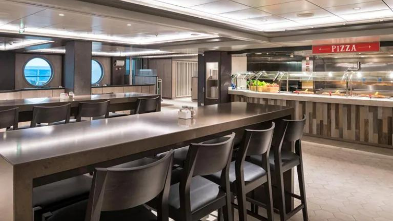 Bufffet do navio MSC Seaside, tendo em primeiro plano, mesas e cadeiras altas e, ao fundo, a estação de pizzas com letreiro vermelho sinalizando o local.