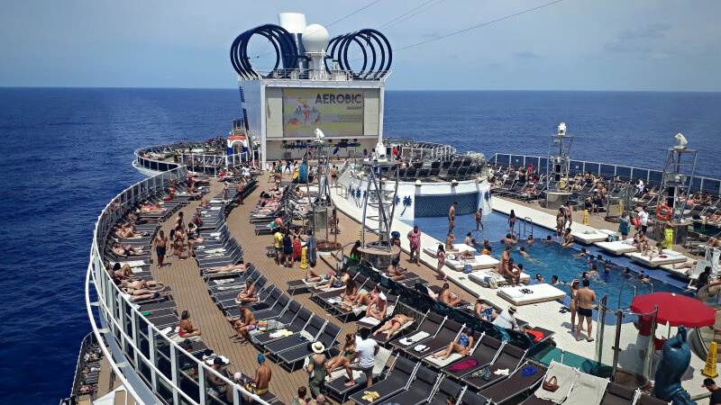 Deck da piscina principal do navio MSC Seaside, contendo várias cadeiras espreguiçadeiras marrons e pessoas tomando sol, tendo o mar ao fundo.