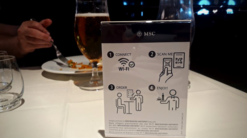Interior do navio MSC Seaside: mesa de restaurante com um material impresso com instruções para leitura de QR-Code impresso para visualização do cardápio.