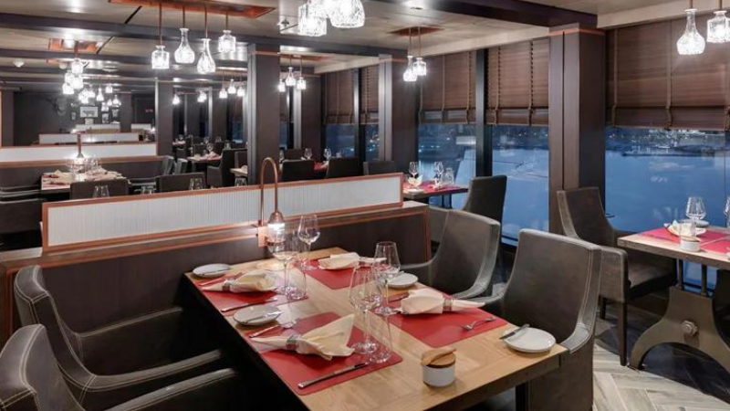 Restaurante Butcher's Curt no navio MSC Seaside, tendo em primeiro plano uma mesa de madeira com jogos americanos vermelhos, guardanapos, copos e talheres, com vista lateral o mar.