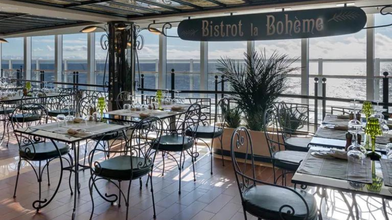 Interior do navio MSC Seaside: ambiente com várias mesas e cadeiras de ferro, com vista para o mar, com placa sinalizando nome do local - Bistrot La Bohème.