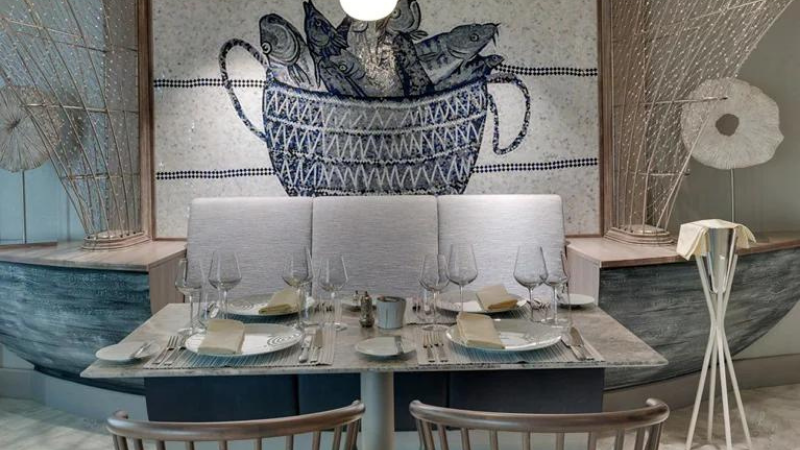 Interior do navio MSC Seaside: mesa de restaurante arrumada com pratos, copos e talheres, num ambiente com decoração em tons azulados e brancos, tendo ao fundo uma parede com quadro de cesta contendo peixes.