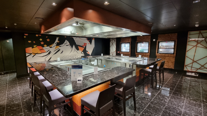 Balcão do Restaurante Teppanyaki, com balcão quadrado de pedra marrom e cadeiras altas marrons, dispostas em frente ao mesmo.