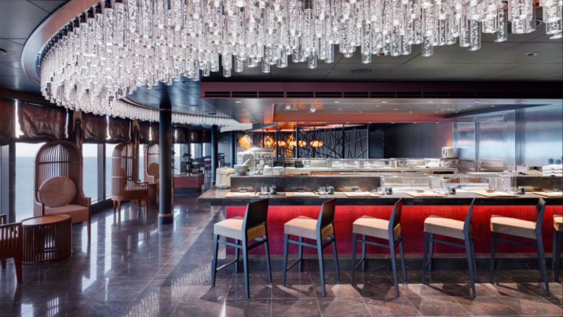 Balcão de sushi, com parede vermelha e cadeiras altas em frente ao mesmo, e teto decorado com pingentes de vidro.