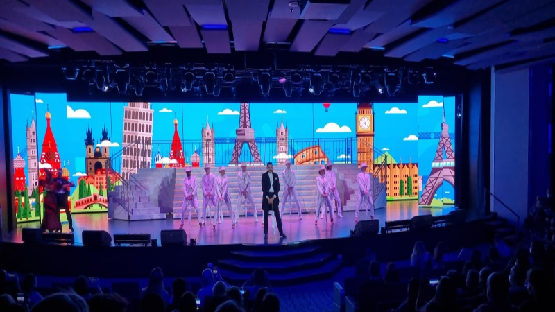 Palco de teatro do navio MSC Seaside, com cenografia de fundo de palco com impressão da Torre Eifel, Torre de Piza, igrejas e edifícios, tendo a frente um grupo de dançarinos vestidos de fraque branco e um apresentador com fraque preto.