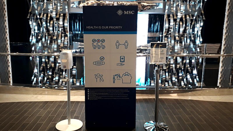 Totem de sinalização preto com impressão de regras de segurança: uso de máscara, distanciamento social e lavagem das mãos. Ao lado, um totem contendo álcool gel.